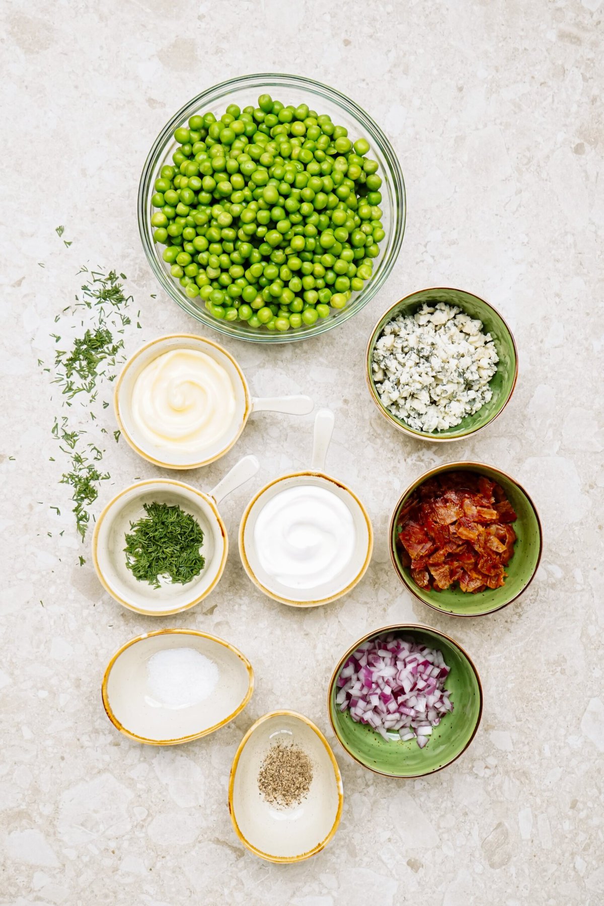 Assorted ingredients for a pea salad recipe arranged neatly on a light surface, including green peas, herbs, dairy products, and seasonings.