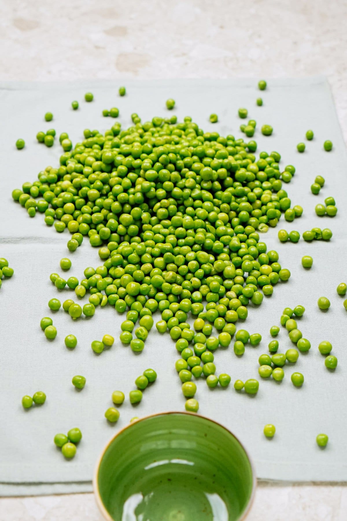 A pile of fresh green peas intended for a pea salad scattered on a light-colored surface with an overturned green bowl to the side.