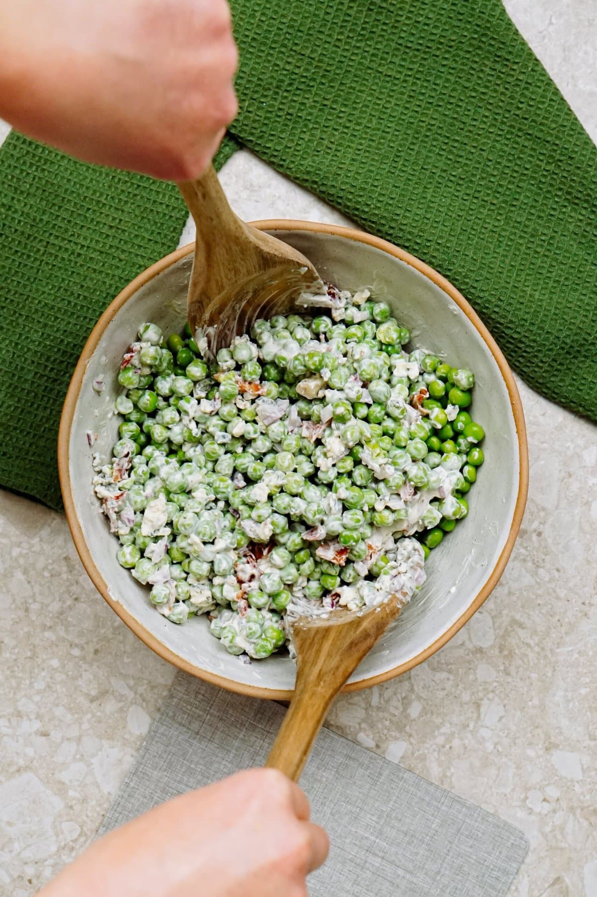 Hands mixing a pea salad with wooden spoons in a bowl.