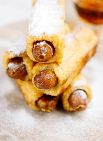 A stack of breakfast pigs in a blanket with powdered sugar on a plate.