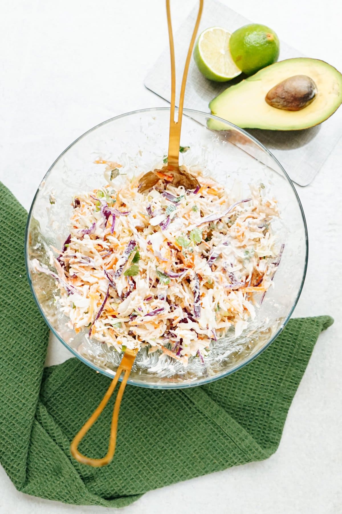 A glass bowl of coleslaw with serving utensils, accompanied by lime slices and an avocado on a kitchen countertop, ready to garnish fish tacos.