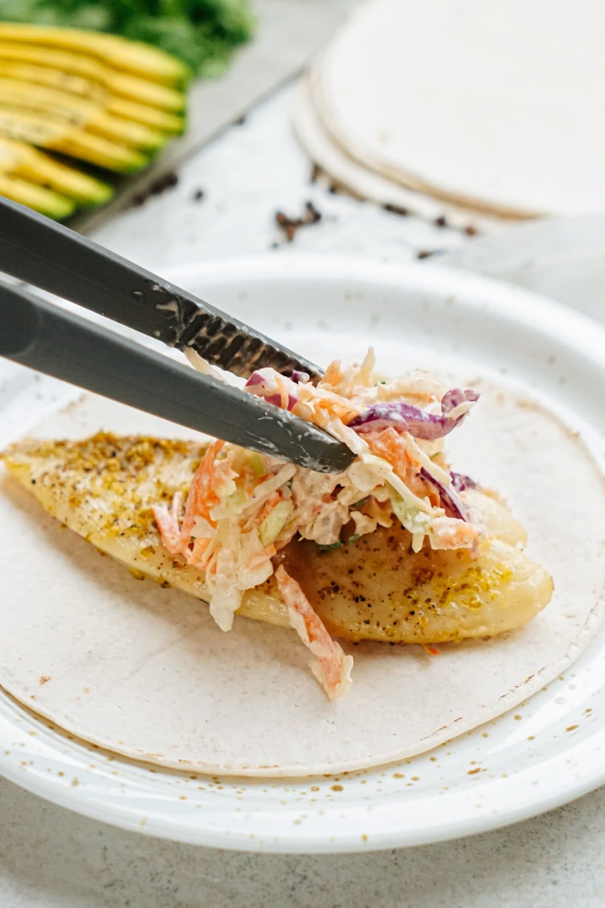 Tongs placing coleslaw on top of a seasoned, grilled fish taco fillet on a plate.