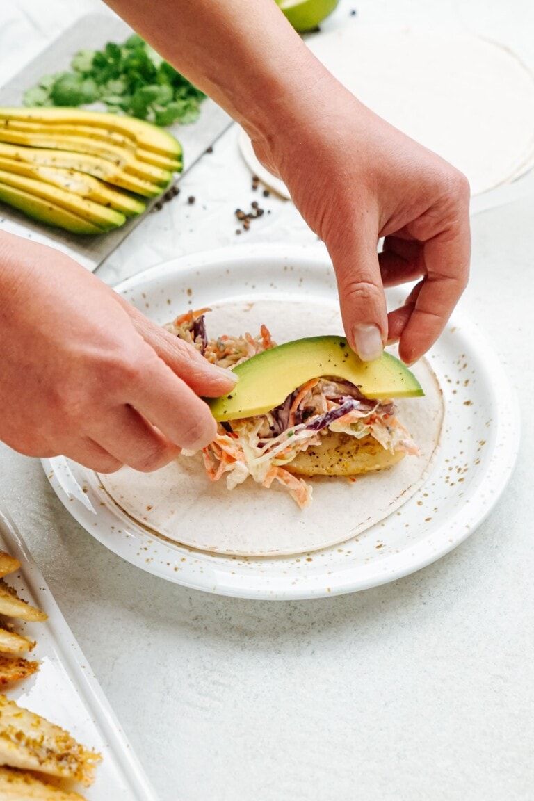 Person adding avocado slices to fish tacos on a white plate.