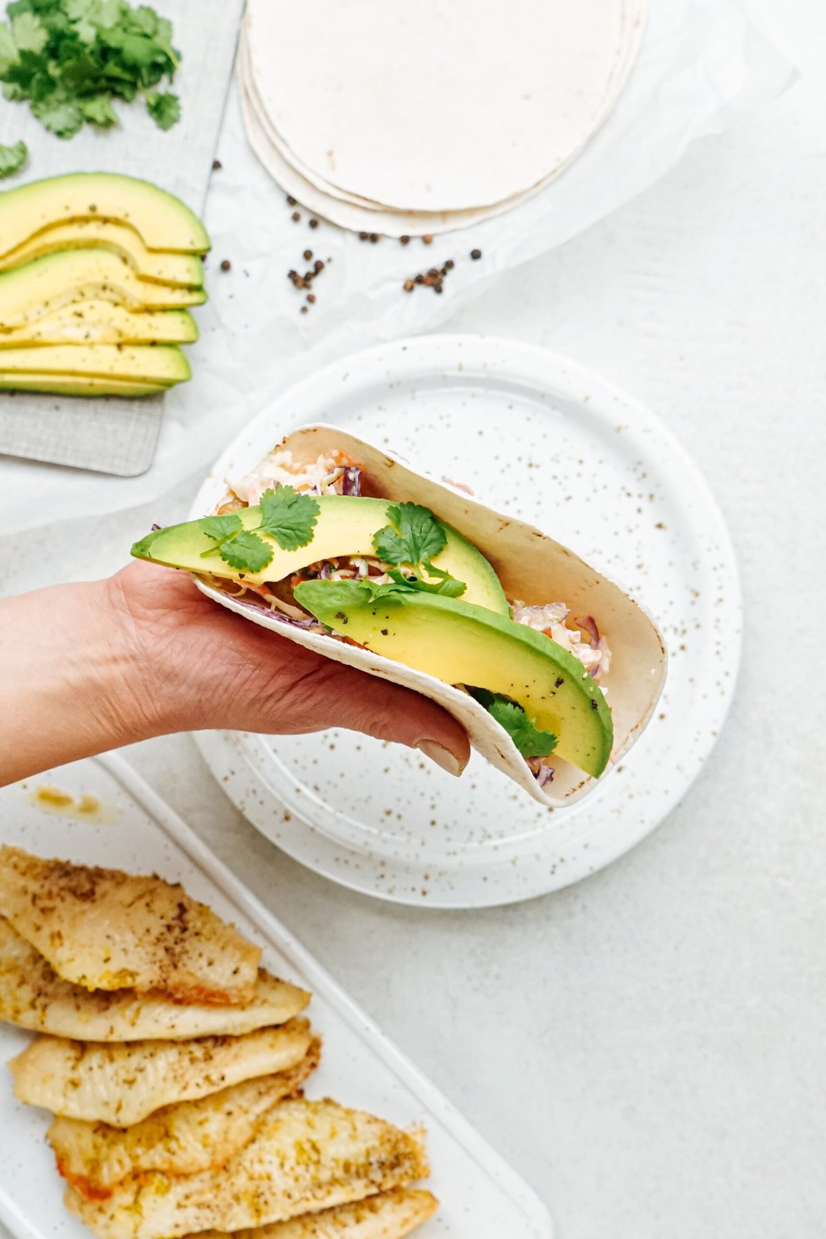 A hand holding a fish taco filled with avocado and topped with cilantro, served on a speckled plate alongside grilled chicken strips.