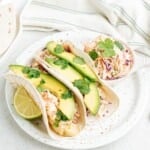 Two fish tacos with avocado and lime on a plate, garnished with cilantro.