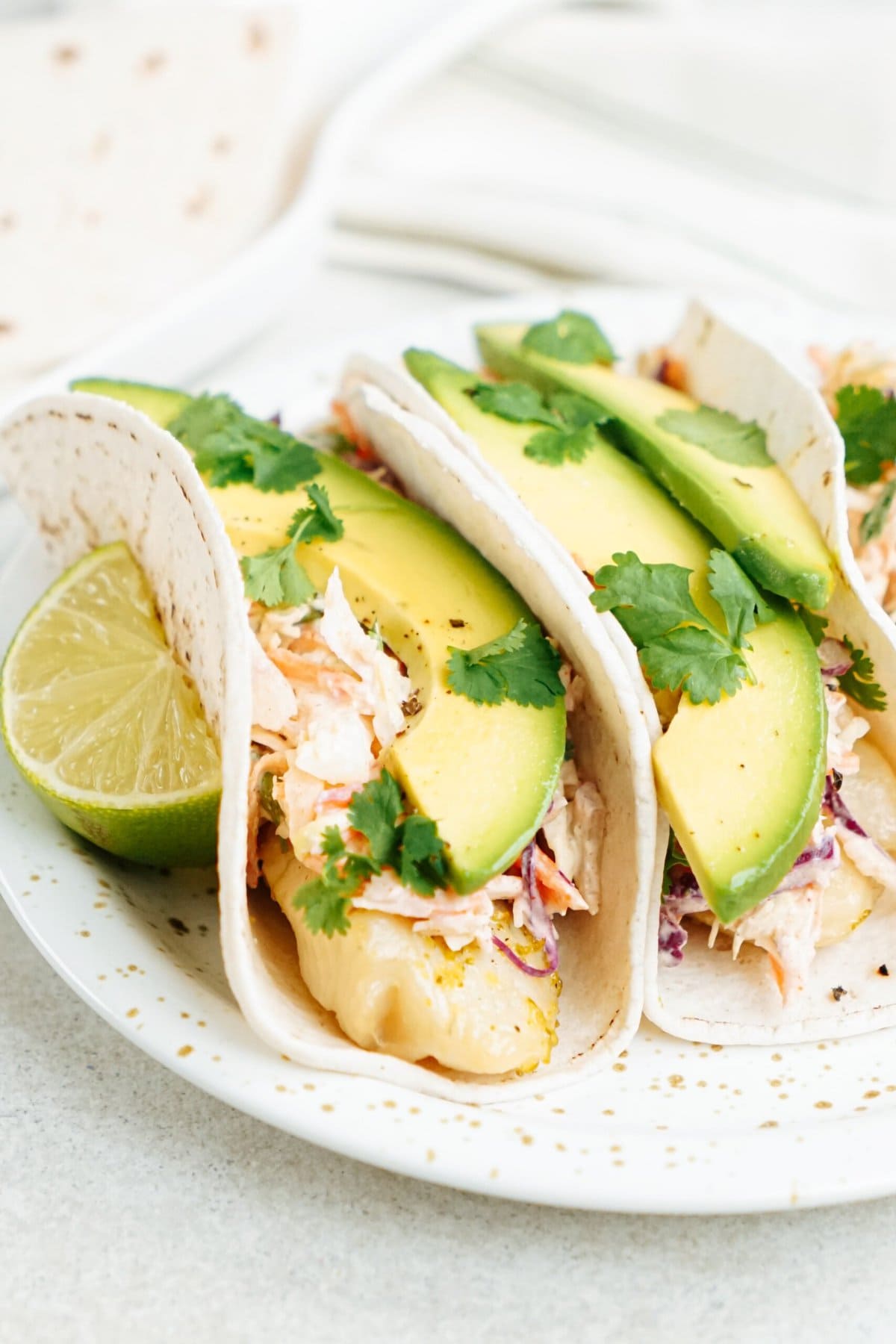 Three fish tacos with avocado and lime on a plate.