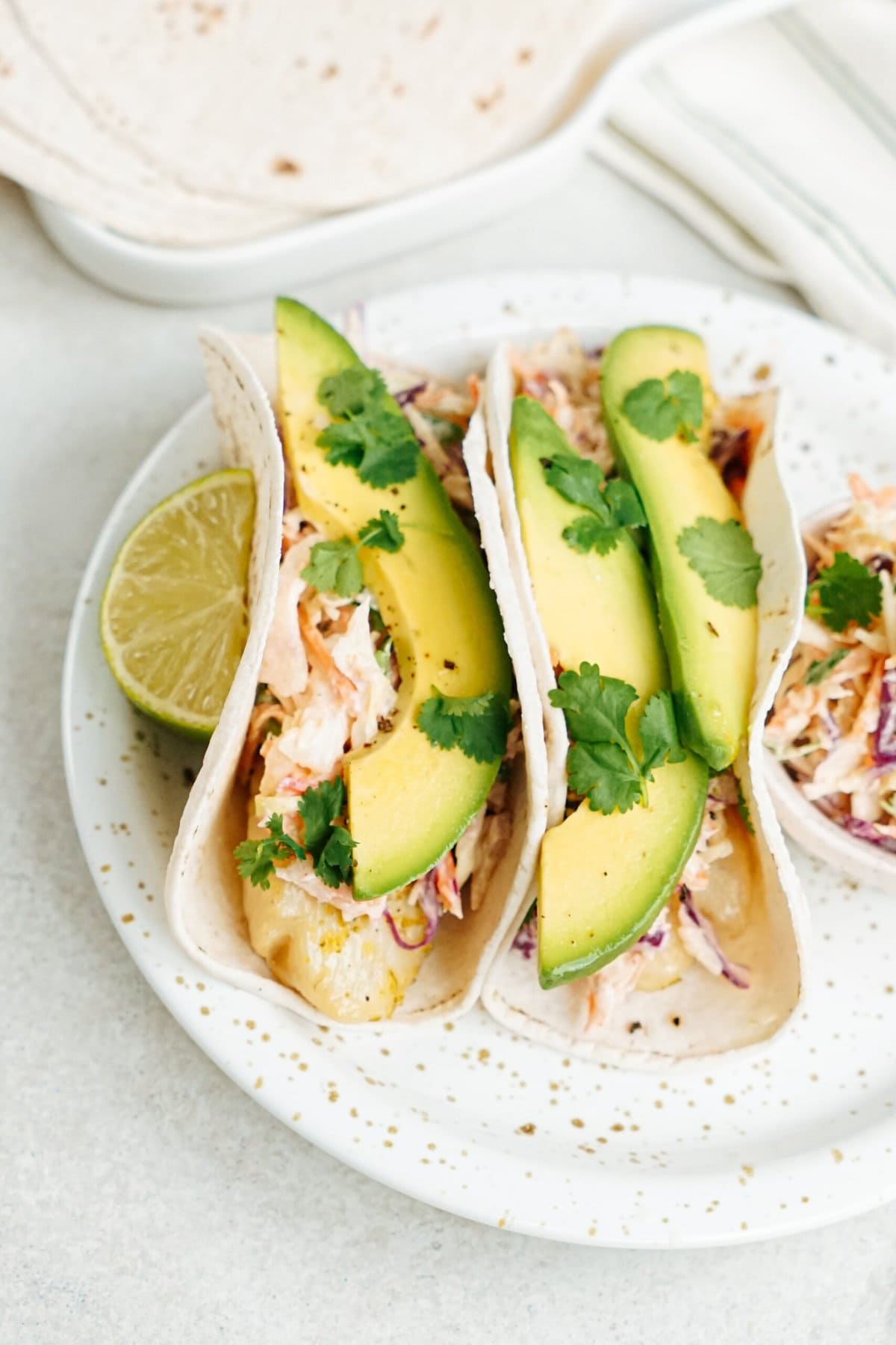 Three fish tacos garnished with avocado slices and cilantro, served with a lime wedge on the side.