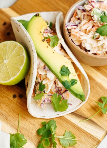 A fresh fish taco with avocado and coleslaw, accompanied by a wedge of lime and cilantro.