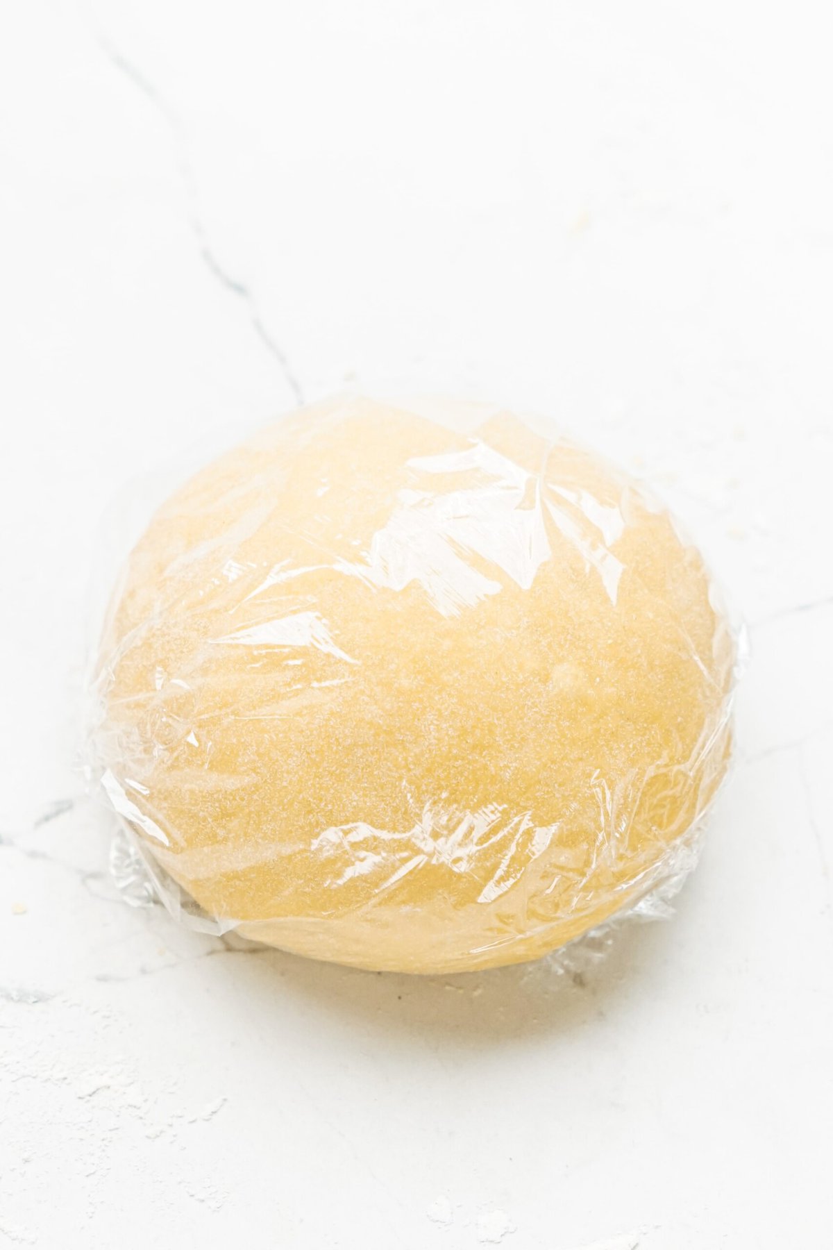 A ball of dough wrapped in plastic on a white surface.