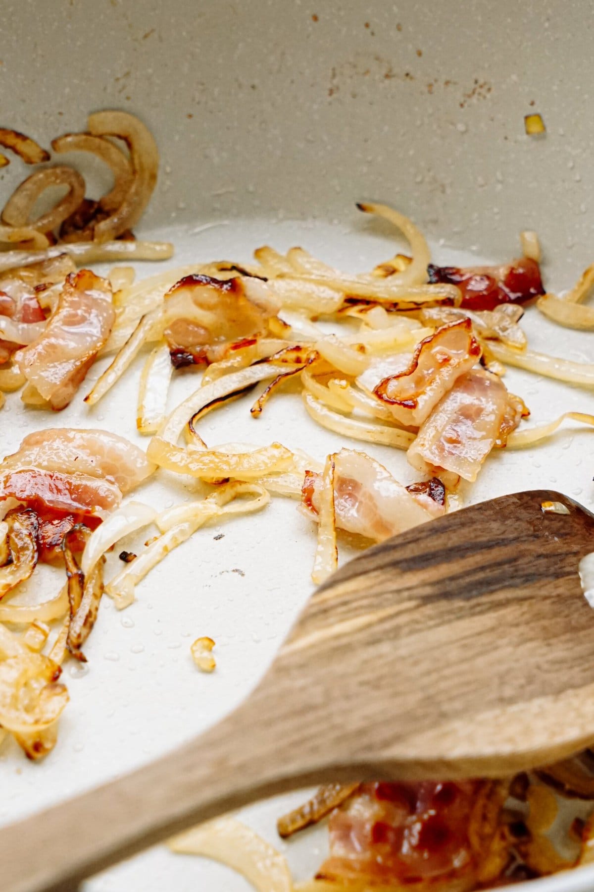 Bacon and onions in a pan with a wooden spoon.