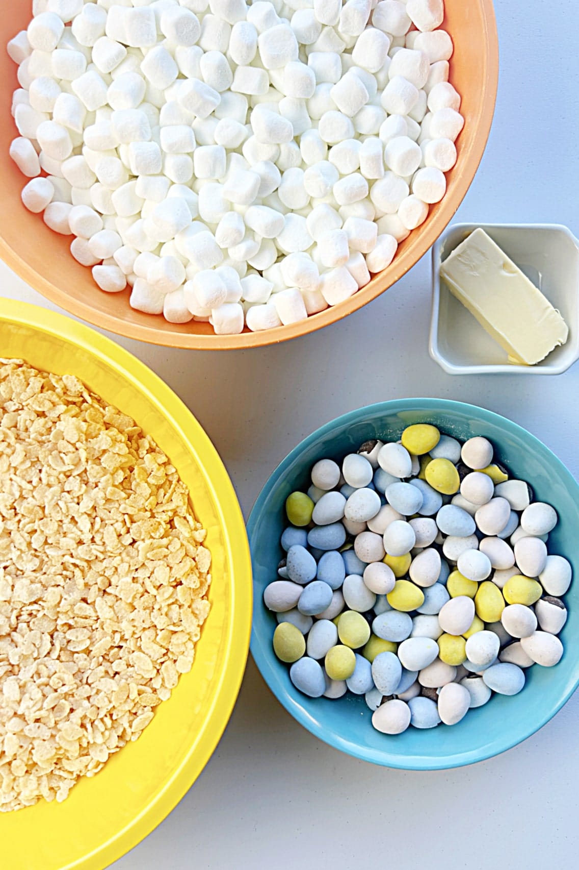 Assorted ingredients for Rice Krispie Treats including marshmallows, chocolate candies, crisped rice cereal, and butter displayed in separate colorful bowls.