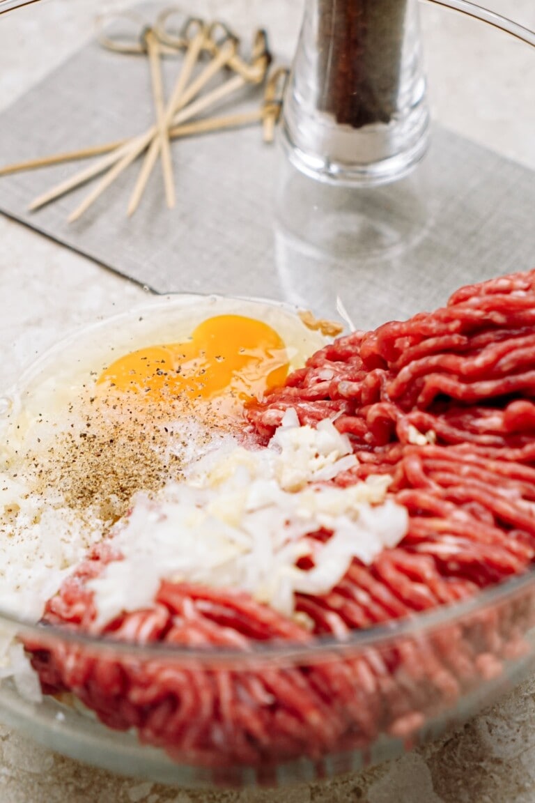 Ingredients for meat preparation: ground beef, an egg, and spices in a glass bowl.