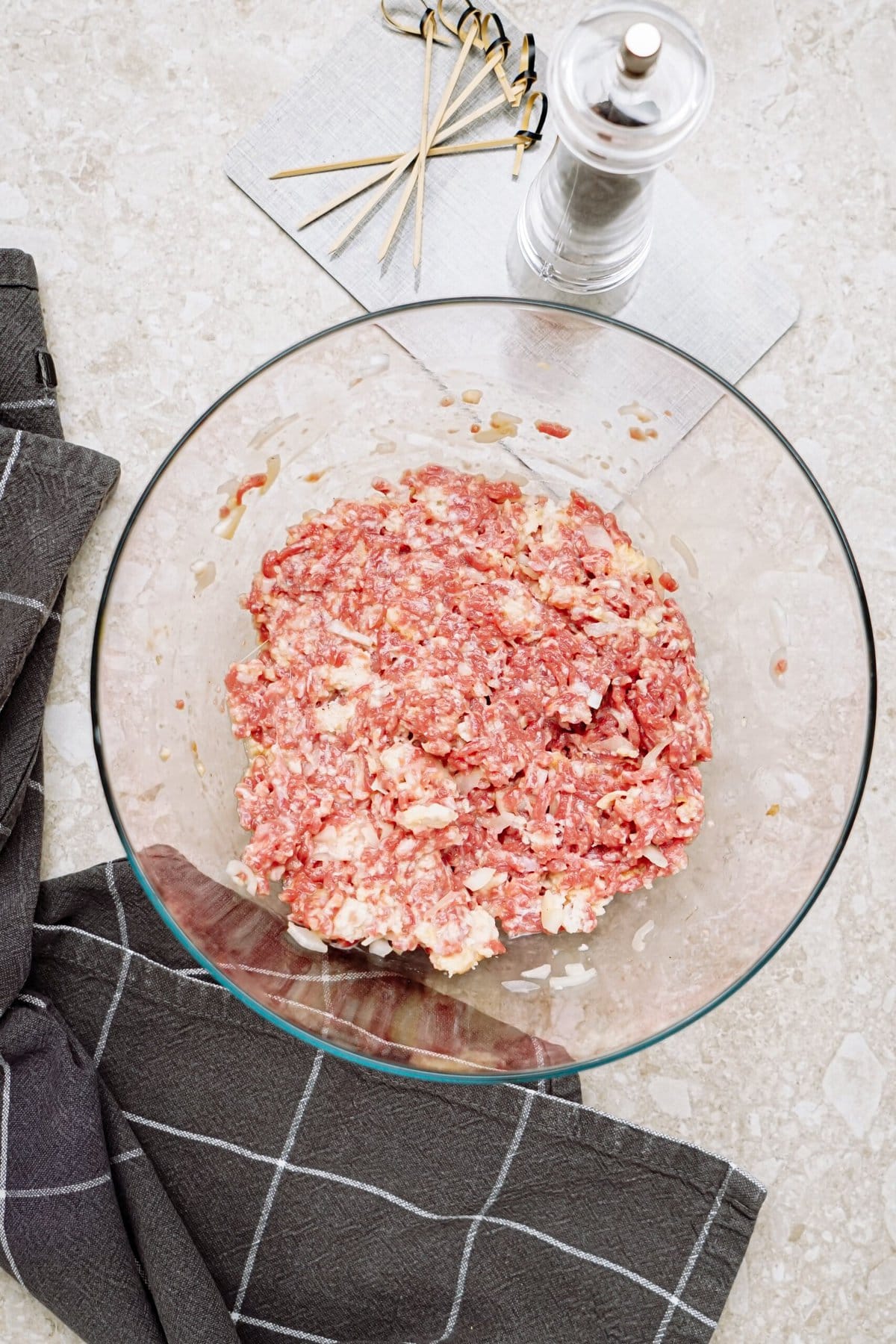 Ground meat mixture in a glass bowl with kitchen utensils and seasoning nearby.