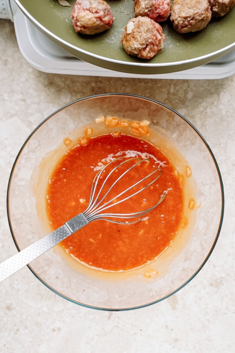 Mixing sauce with a whisk in a glass bowl next to a plate of meatballs.