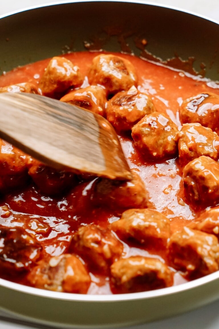 Meatballs simmering in sweet and sour sauce in a cooking pan with a wooden spoon.
