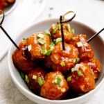 A bowl of glazed meatballs garnished with green onions, served with toothpick skewers.