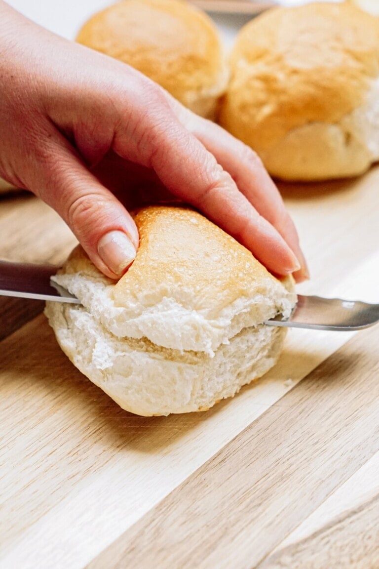 A hand slicing a bread roll with a knife on a wooden cutting board.