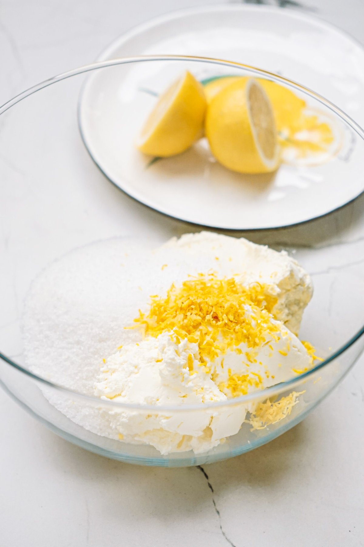 Bowl of ingredients including sugar, cream, and lemon zest, with lemon wedges on a plate in the background.