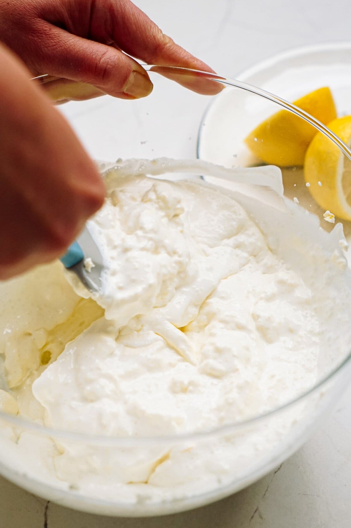 A person stiring white cream from a glass bowl with sliced lemons in the background.