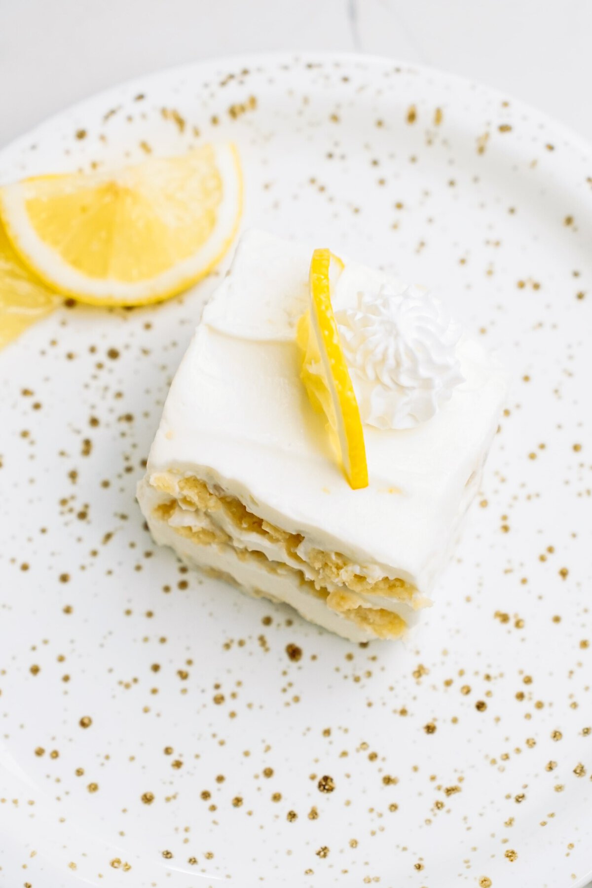 A slice of lemon cake with white frosting, garnished with a lemon peel and whipped cream on a white plate sprinkled with gold-colored sugar.
