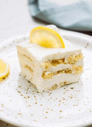 A slice of lemon dessert with creamy layers on a plate, garnished with a lemon slice and zest.
