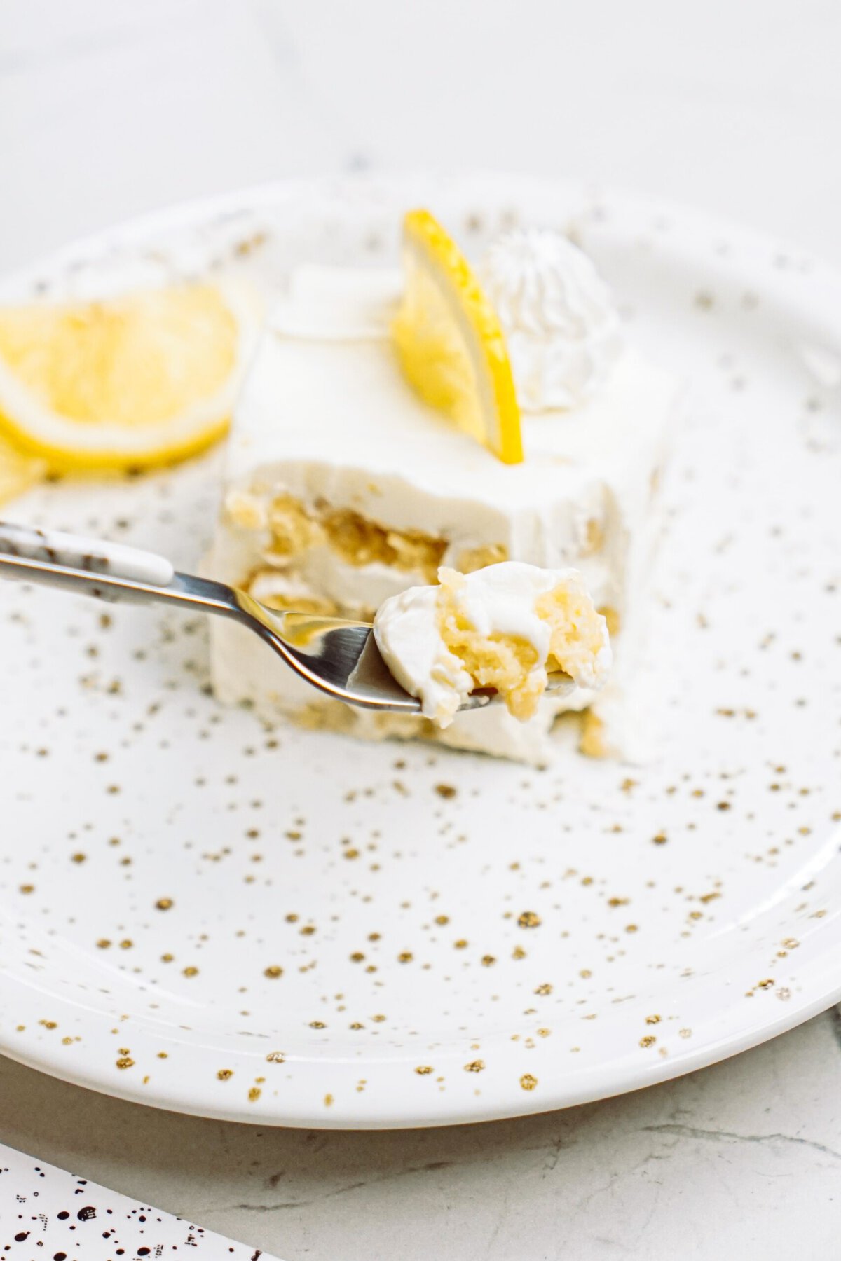 A slice of lemon cake with whipped cream on a speckled plate, garnished with a lemon slice, with a fork taking a piece.