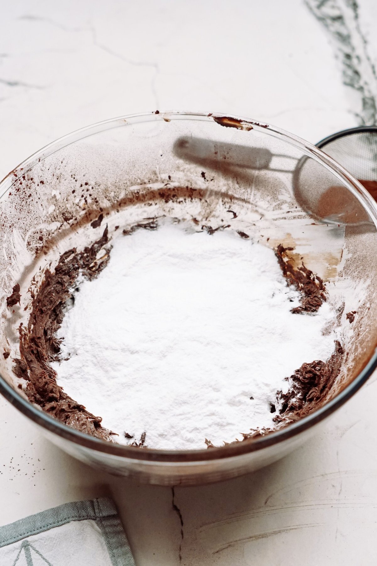 A glass bowl containing chocolate frosting with a layer of powdered sugar on top.