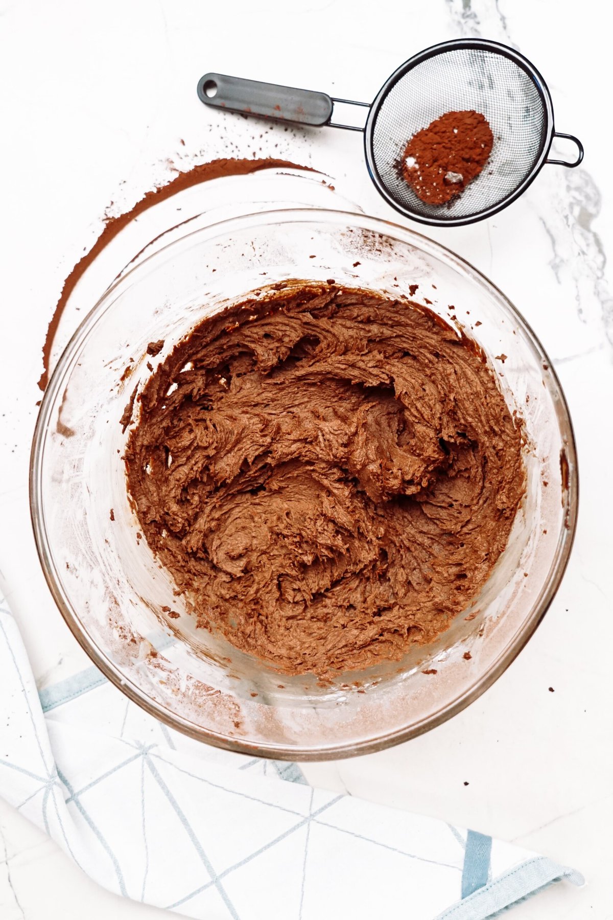 Bowl of chocolate frosting with spilled cocoa powder and sieve on a marble countertop.
