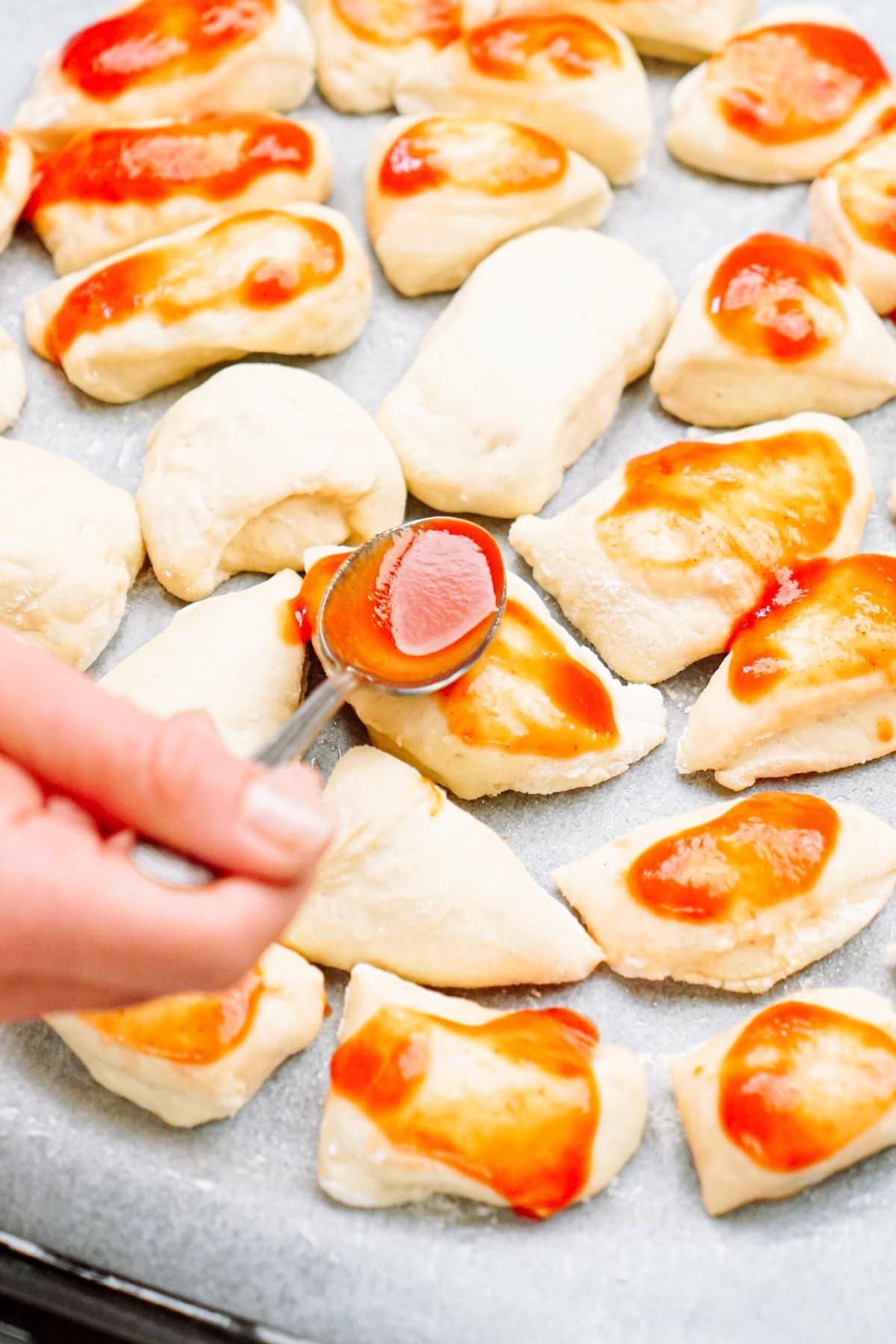 a person spooning pizza sauce onto dough pieces 