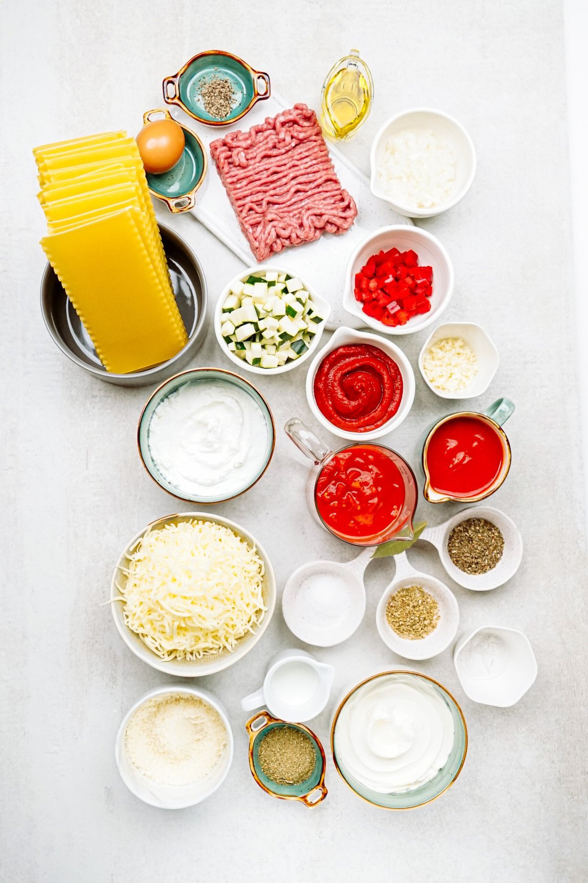 Ingredients for making lasagna laid out, including lasagna sheets, minced meat, eggs, various cheeses, seasonings, and vegetables in bowls and cups on a light-colored surface.