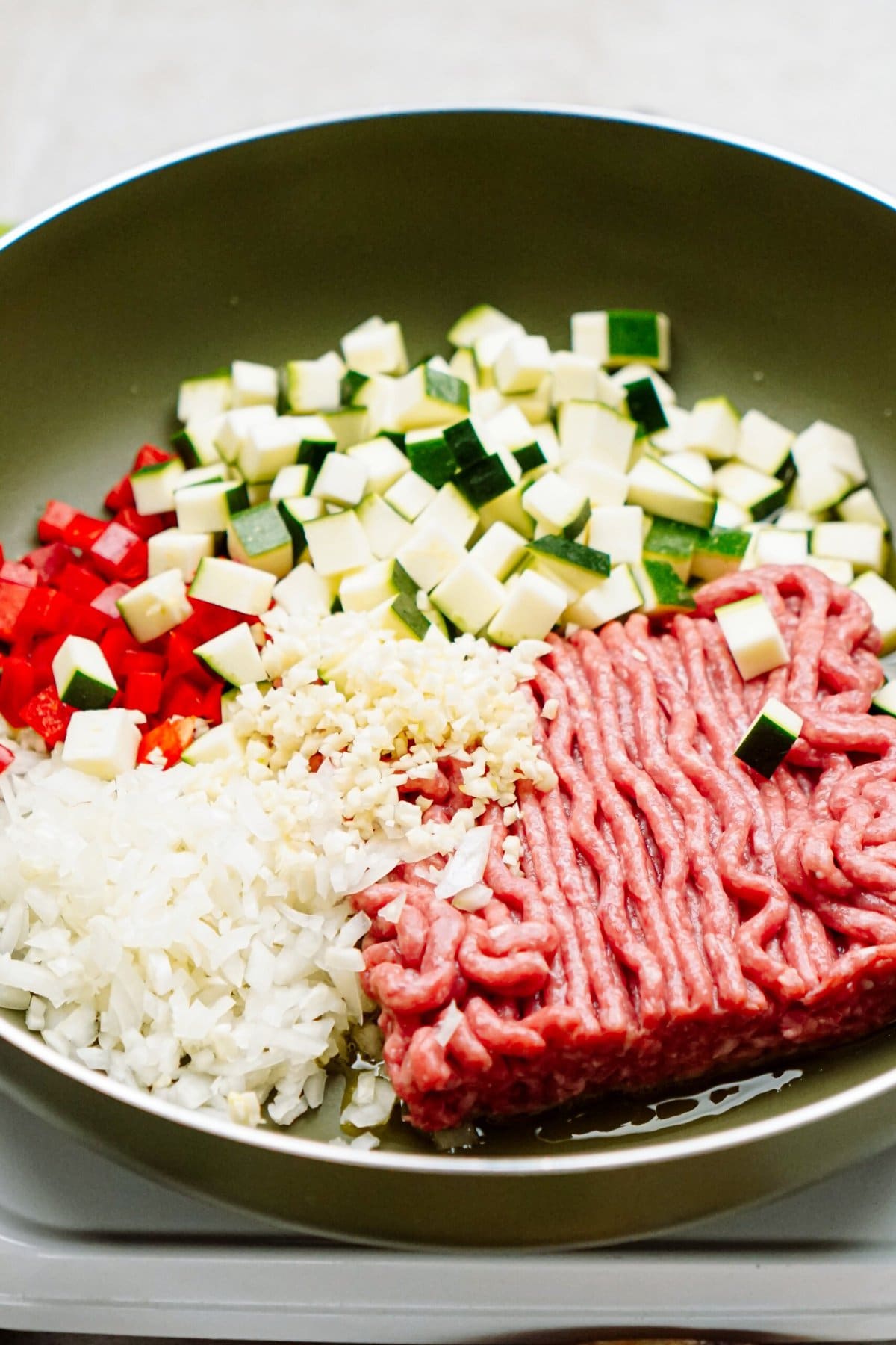 A pan filled with raw ground meat, chopped zucchini, diced red bell pepper, minced garlic, and diced onions.