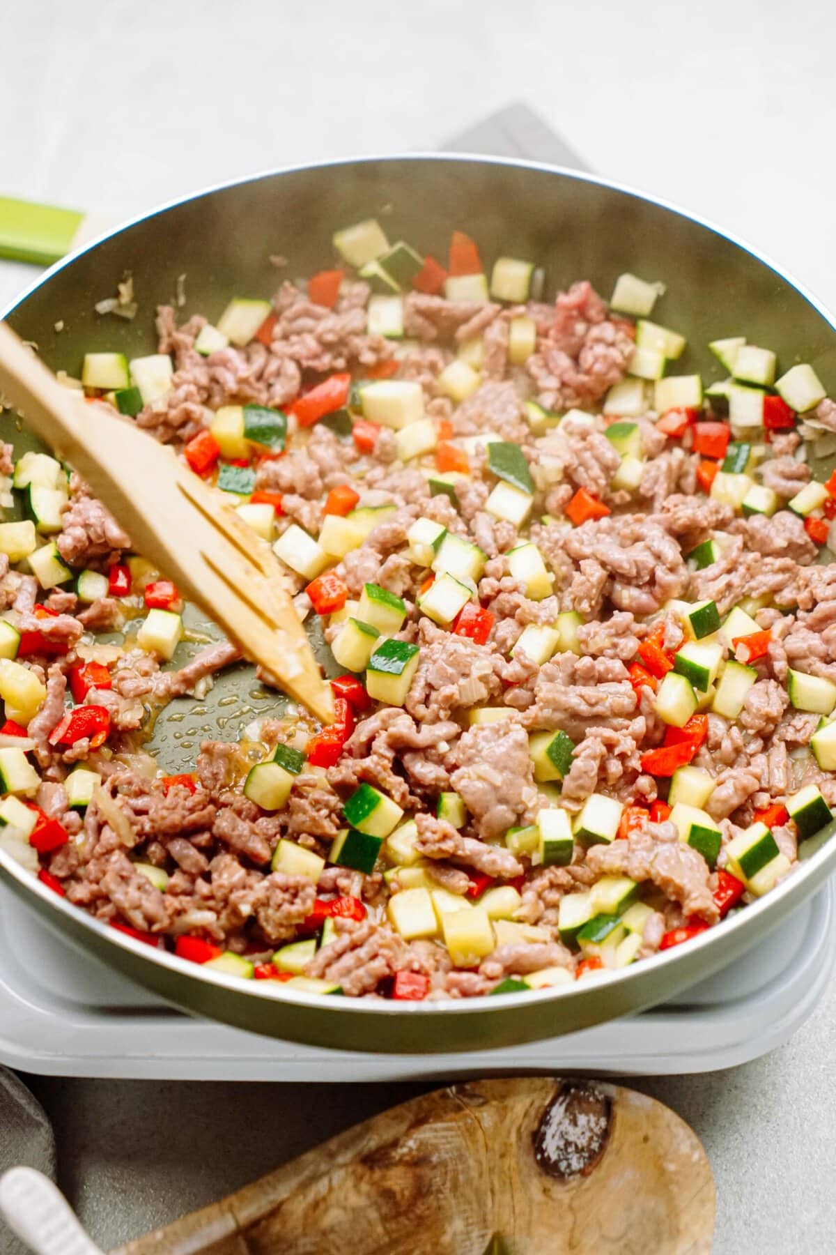 A skillet on a stovetop with minced meat, diced zucchini, red bell peppers, and onions being stirred with a wooden spatula, surrounded by cooking utensils.