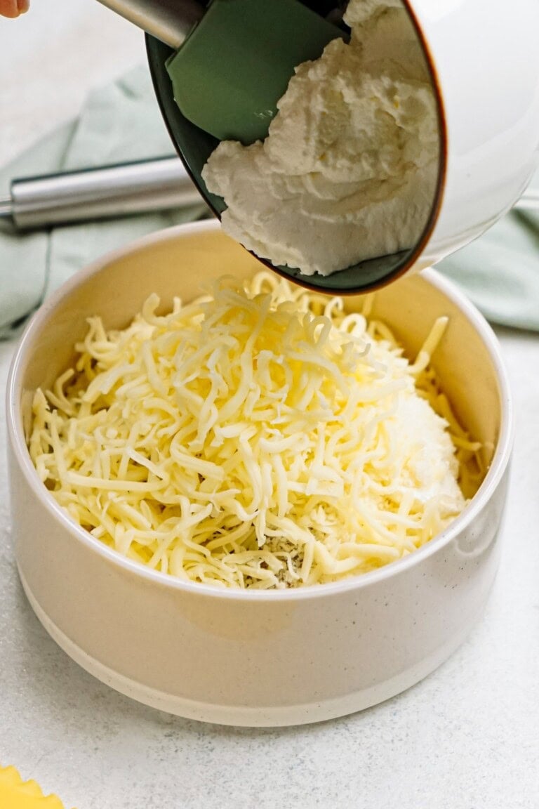 A bowl filled with shredded cheese and a dollop of ricotta being added from a container.