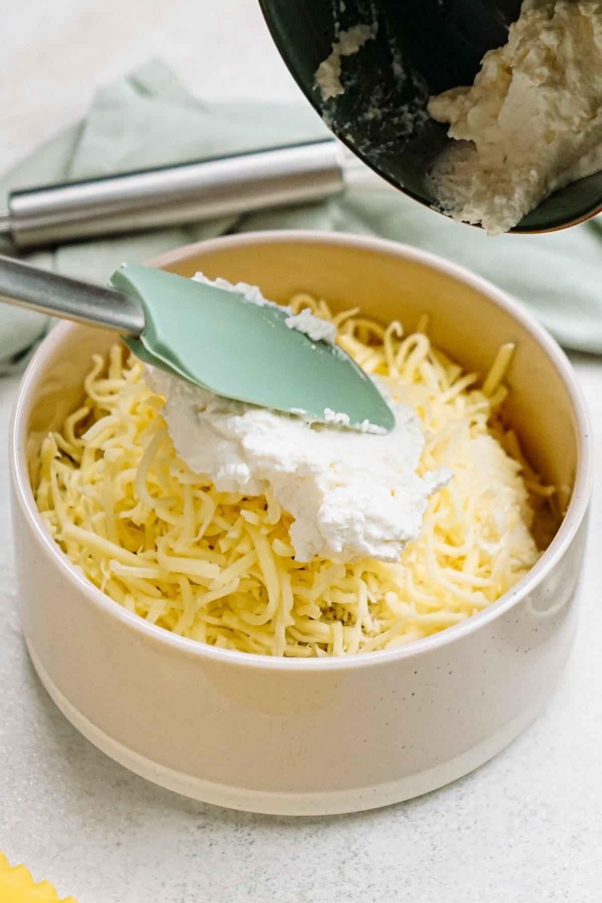 A bowl of shredded cheese is being topped with a dollop of ricotta cheese using a green spatula.