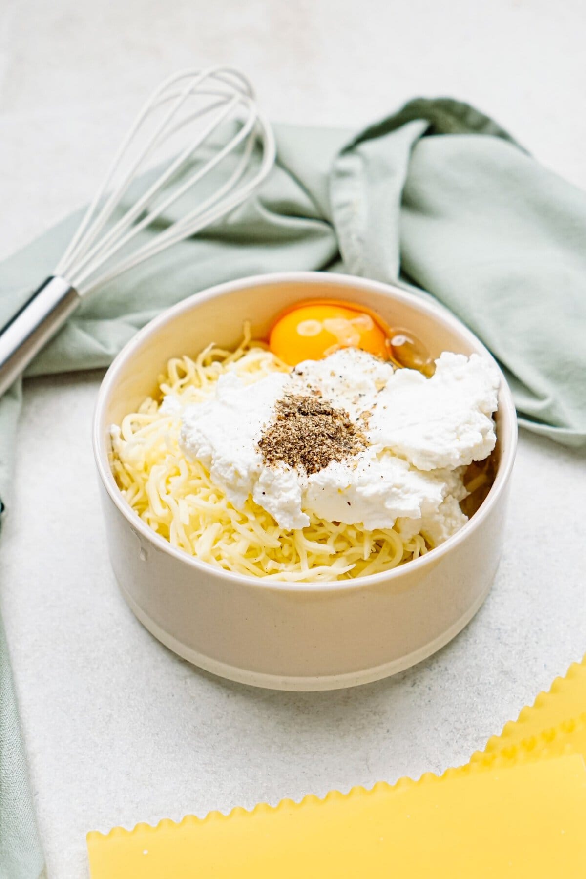 A bowl containing shredded cheese, ricotta, cracked egg, and seasoning sits on a countertop next to a whisk and a green cloth.