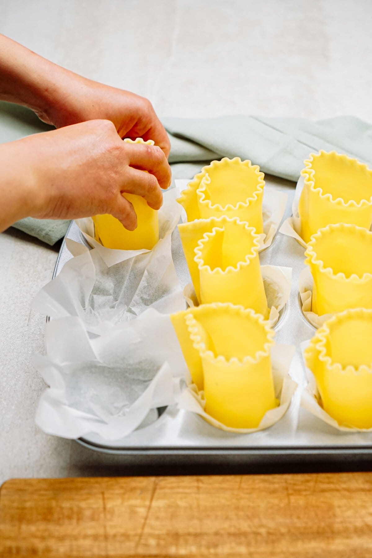 Hands are arranging lasagna noodles rolled into cylindrical shapes in parchment-lined muffin tin compartments on a table.