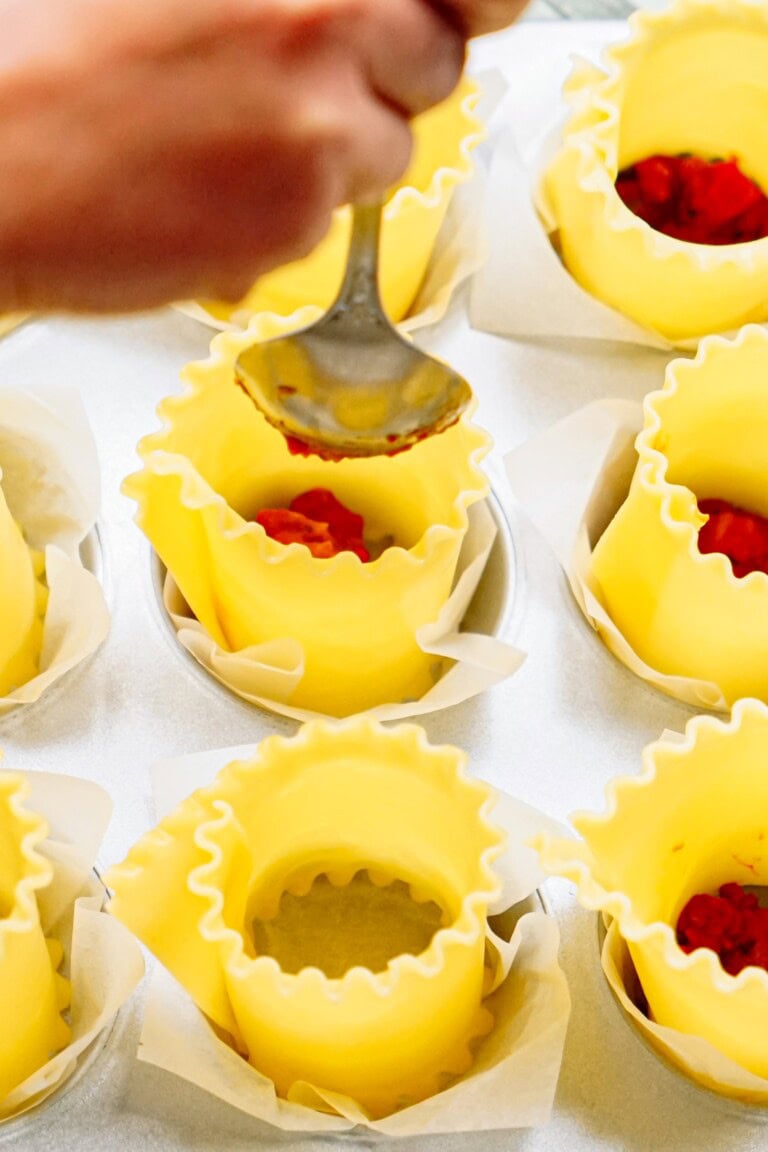 A person is spooning tomato sauce into yellow lasagna noodle cups lined with parchment paper in a muffin tray.