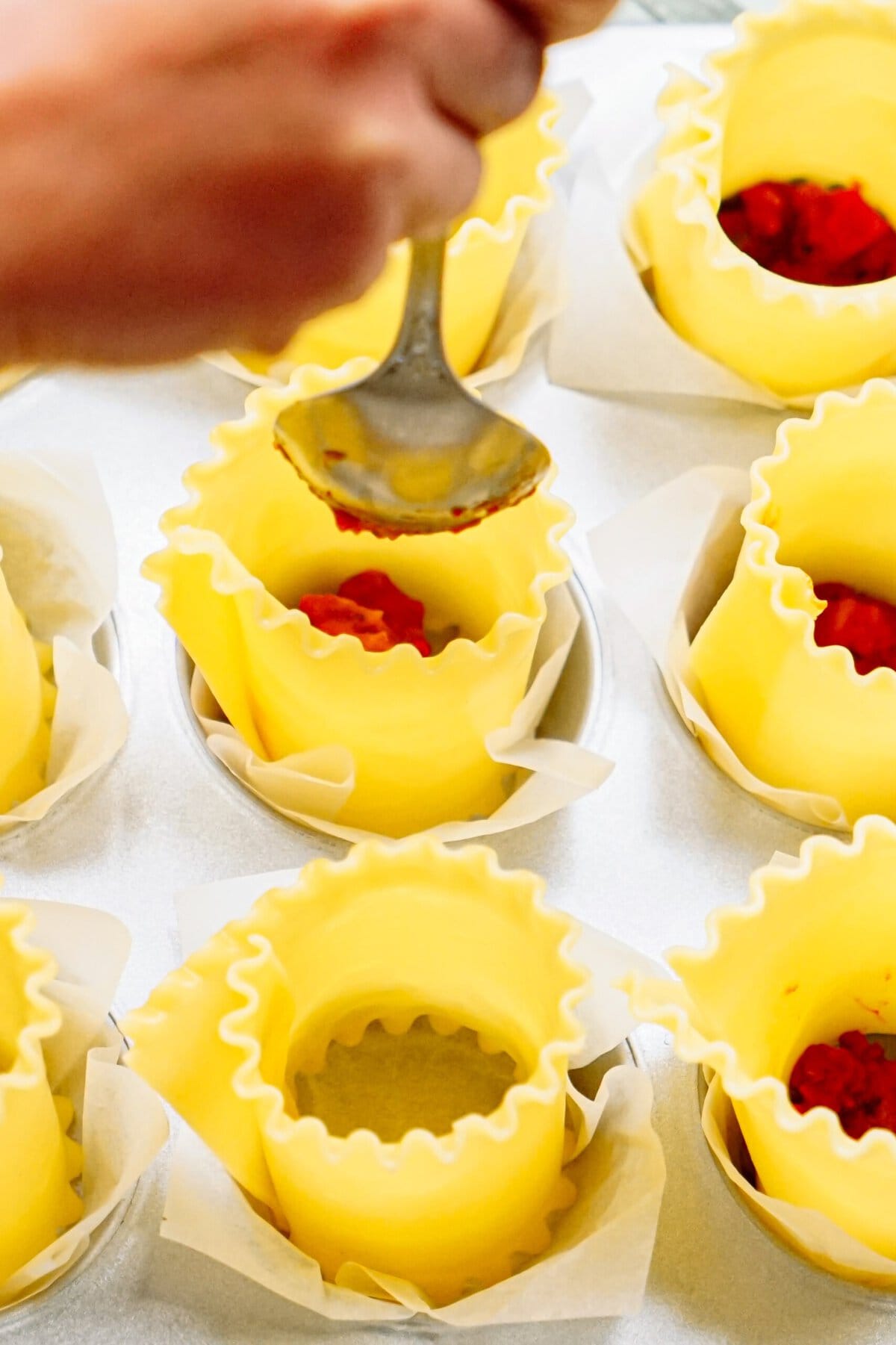 A person is spooning tomato sauce into yellow lasagna noodle cups lined with parchment paper in a muffin tray.