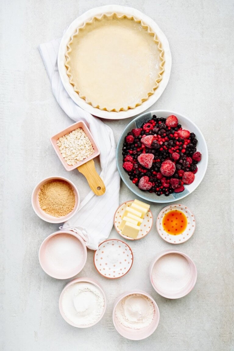 Pie crust in a pan, a bowl of mixed berries, sugar, butter, vanilla, and various other ingredients in small bowls are neatly arranged on a light surface.