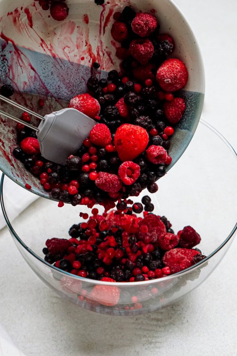 A person transfers a mix of frozen berries including strawberries, raspberries, and blueberries from a white bowl to a clear glass bowl with a spatula.