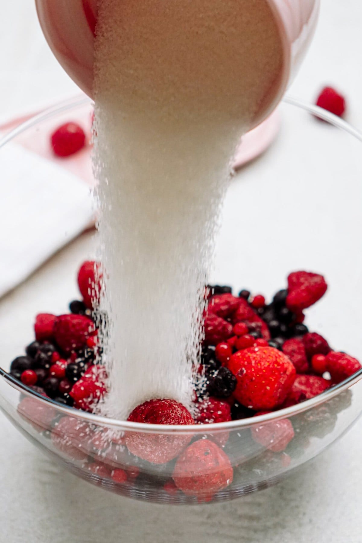 Sugar being poured from a bowl onto a mix of berries in a glass bowl.