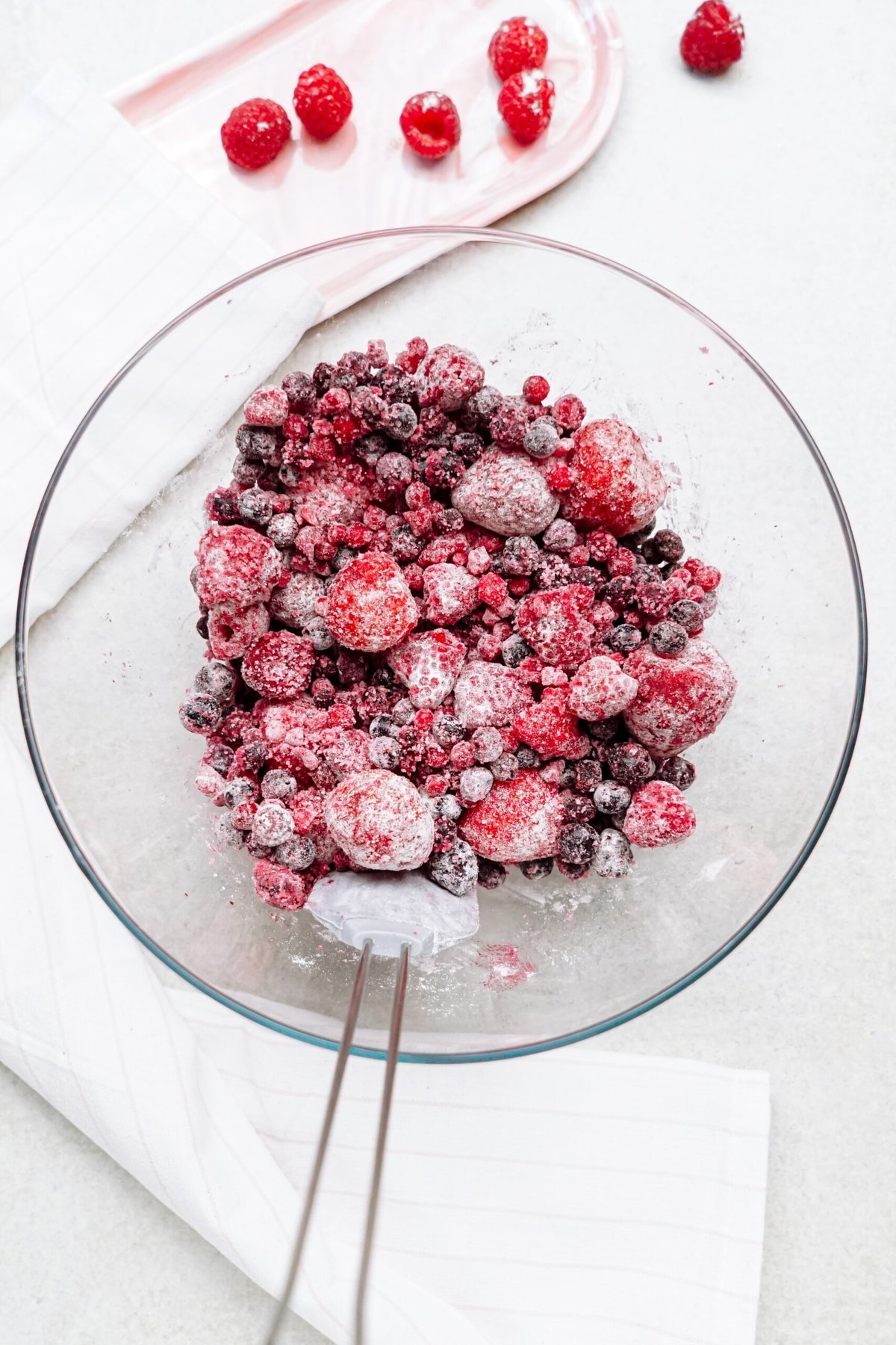 A glass bowl filled with various frozen berries on a white surface, with a few fresh raspberries on a dish in the background.