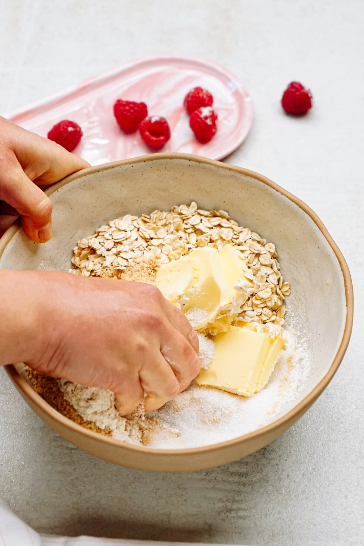 Hands mixing flour, oats, and butter in a bowl. A pink plate with raspberries is in the background.