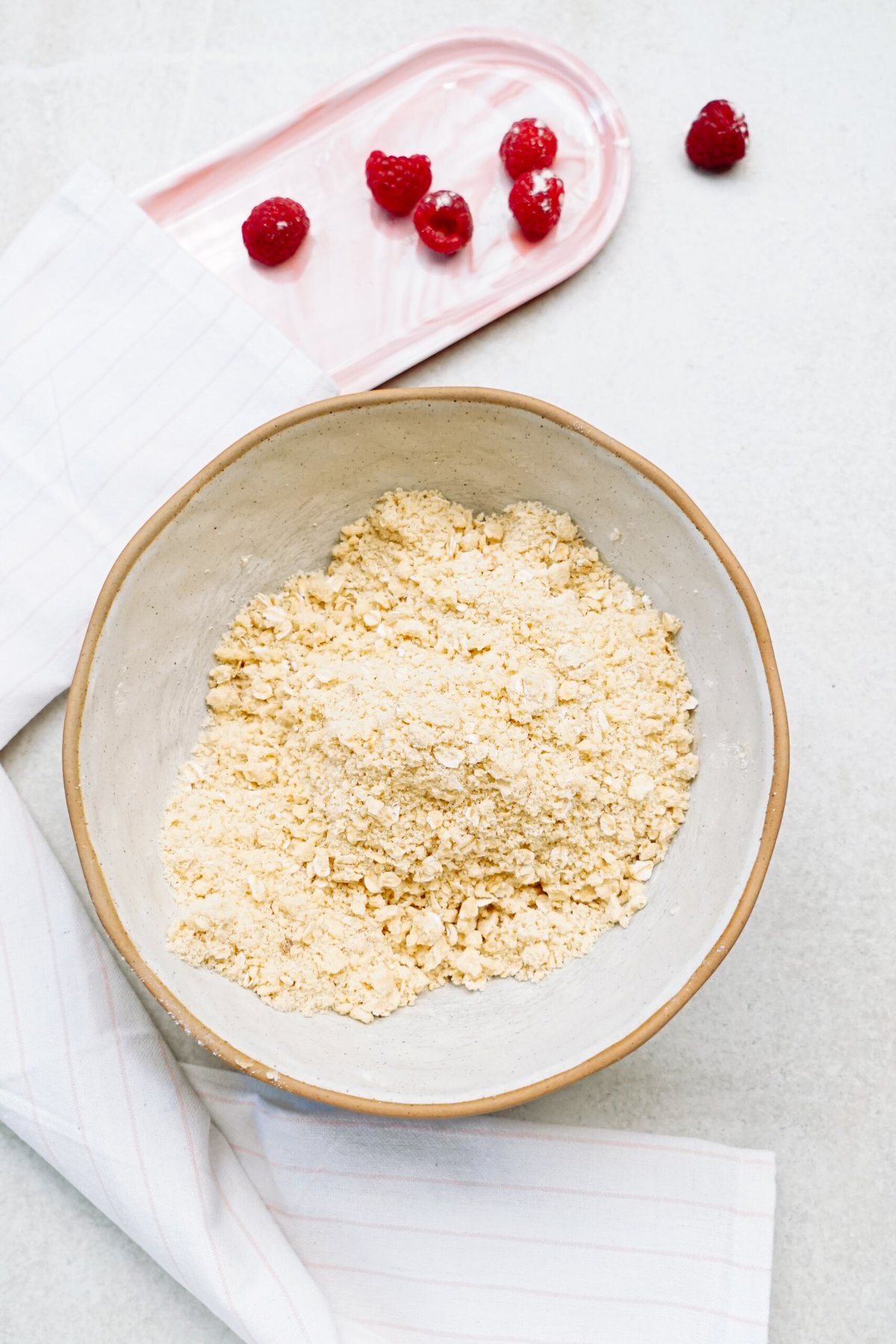 A bowl of crumbled flour with a cloth beside it and a tray of raspberries on the surface.