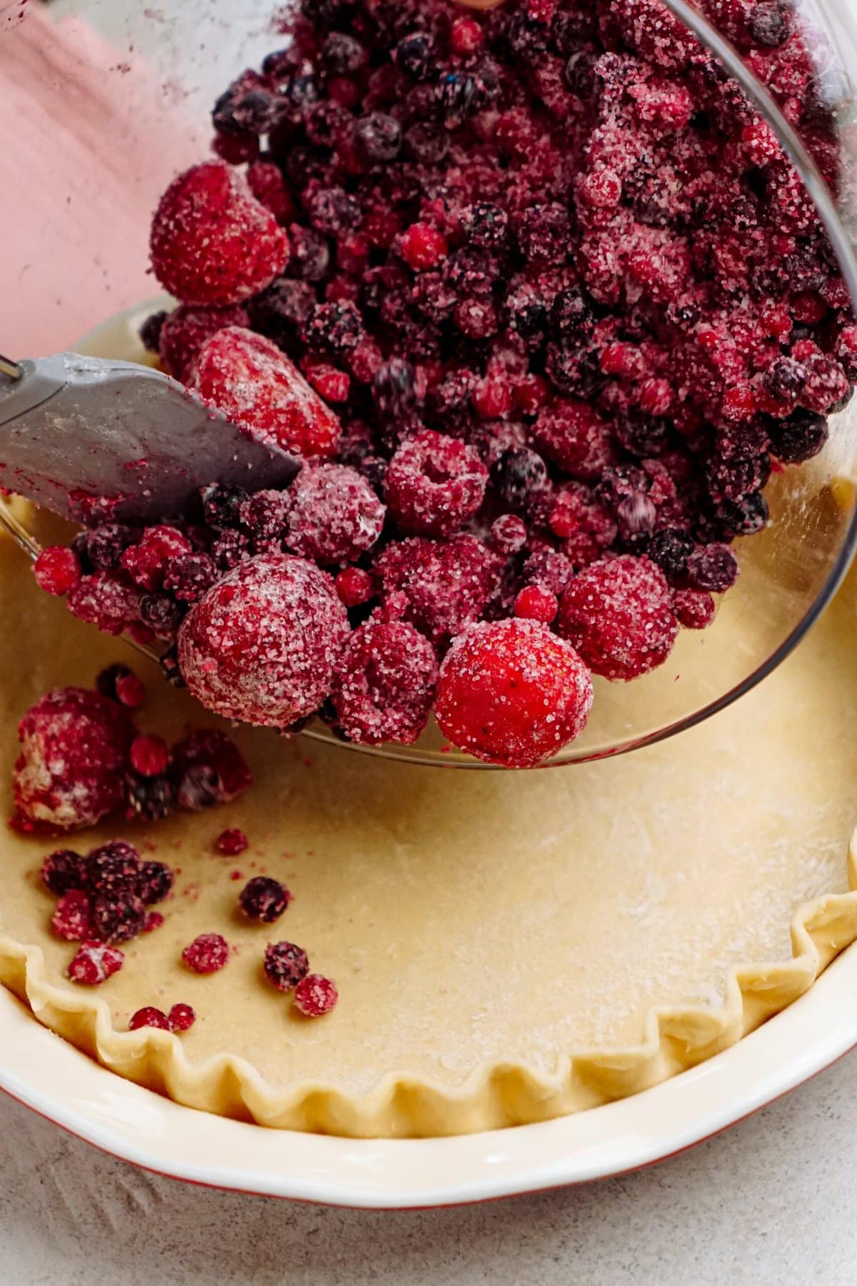 A mixture of frozen berries is being poured into an unbaked pie crust with a fluted edge, preparing to make a fruit pie.