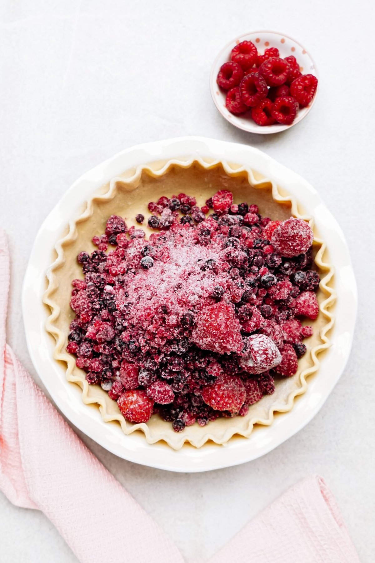 An unbaked pie crust filled with mixed berries and powdered sugar is placed in a white ceramic dish. A pink kitchen towel and a small bowl of raspberries are nearby.