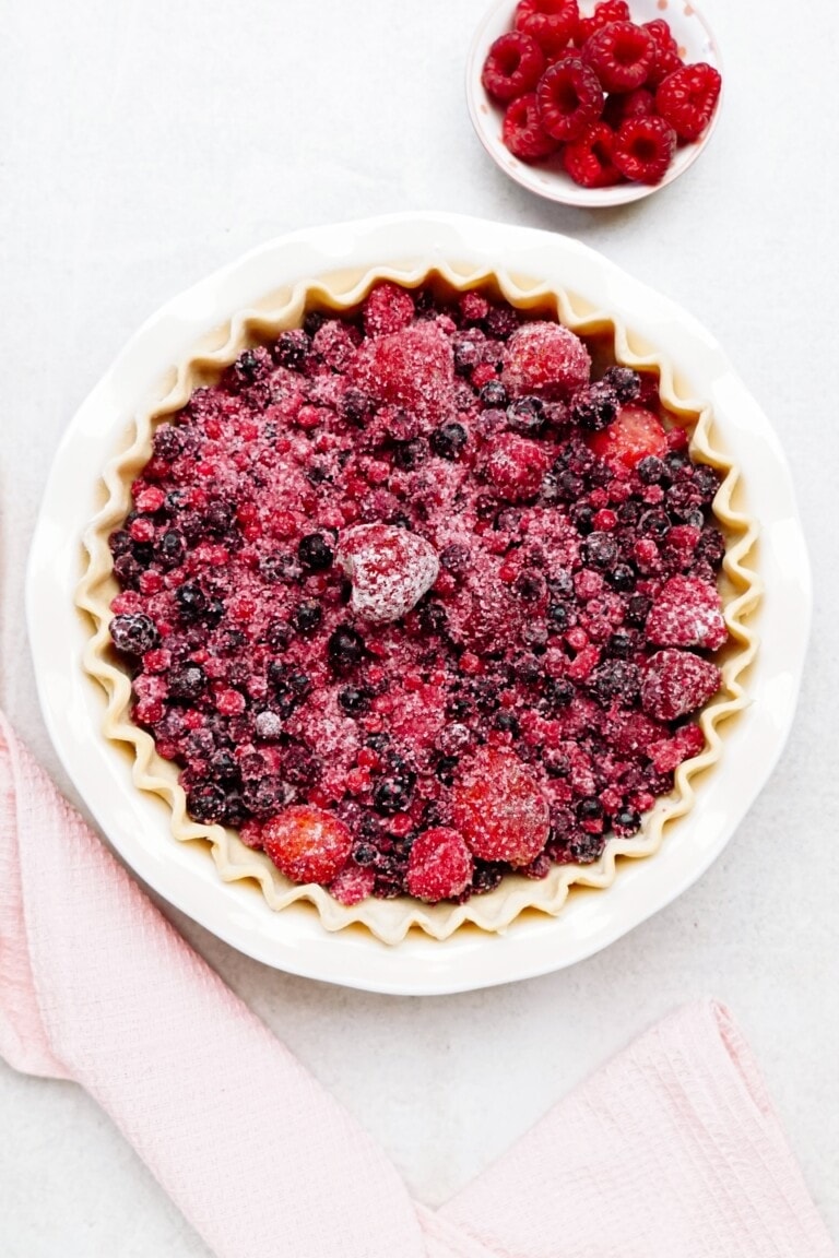 A pie filled with various frozen berries sits on a white surface next to a small bowl of fresh raspberries and a pink cloth.