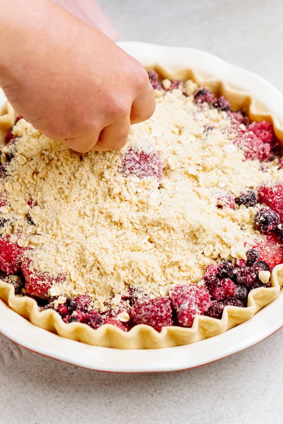 A person sprinkles a crumb topping on a mixed berry pie with a fluted crust, preparing it for baking.