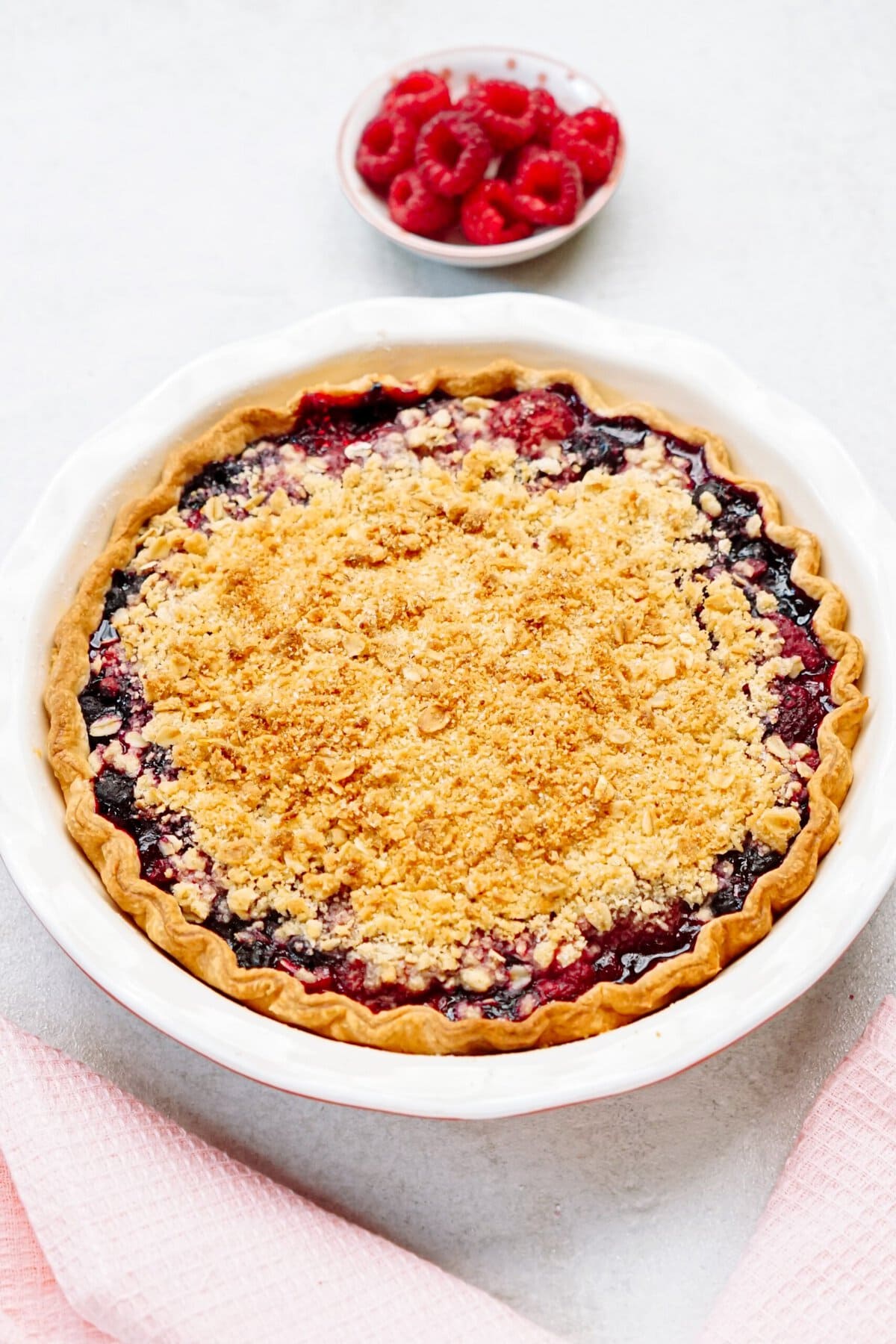 A baked mixed berry streusel pie in a white dish with a small bowl of fresh raspberries in the background.