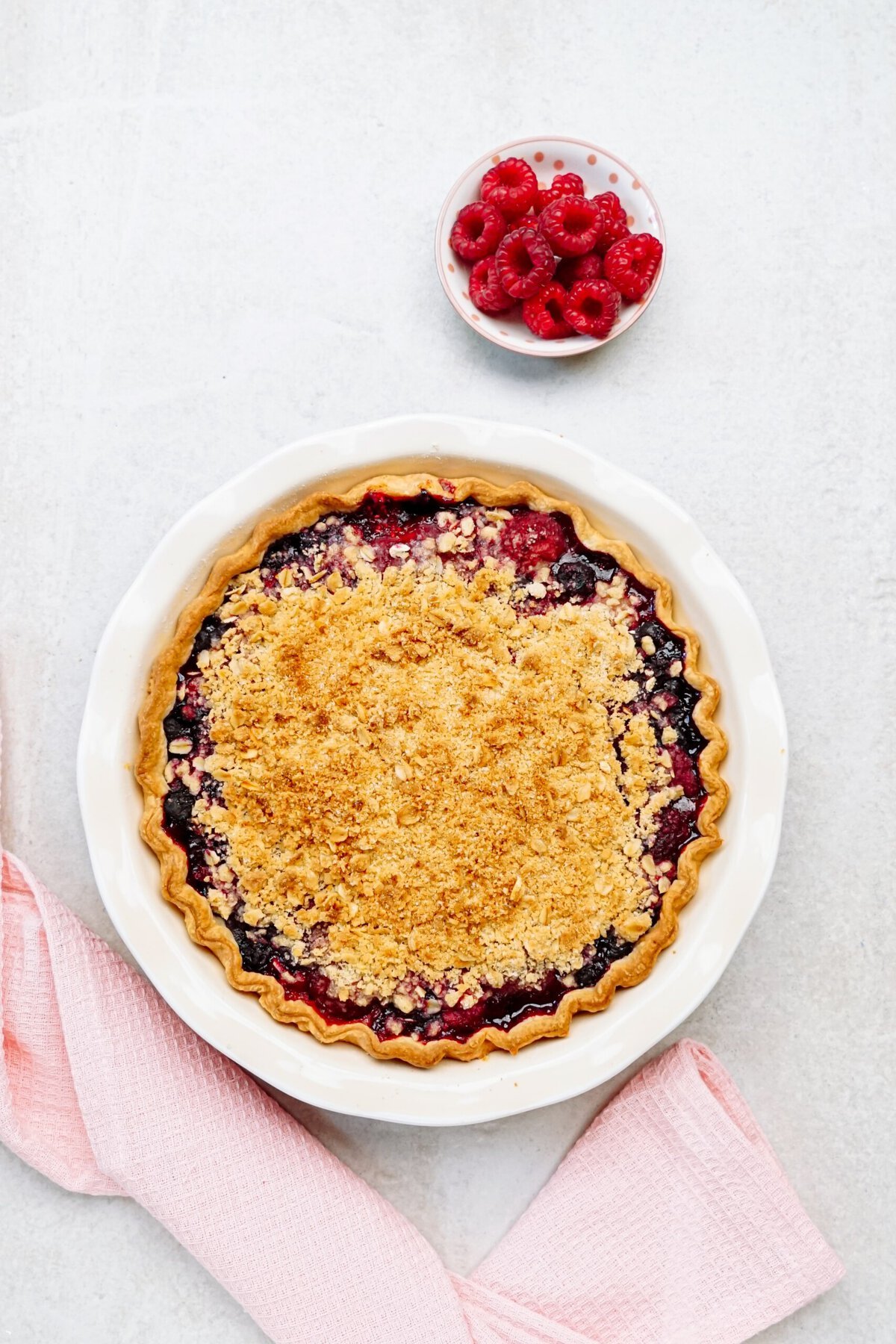 A berry pie with a crumb topping on a white surface. A small bowl of fresh raspberries and a pink cloth are nearby.